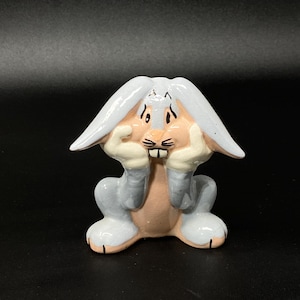 May include: A ceramic figurine of Bugs Bunny, the cartoon character, in a seated position with his hands on his face. The figurine is primarily gray, with pink and white accents. The figurine has a surprised expression.