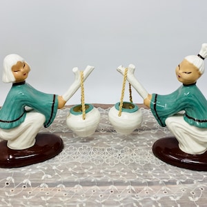 May include: Pair of ceramic figurines depicting figures in teal robes and white pants, each holding a white pot with a gold-colored rope. The figurines are on brown oval bases, with white hair and closed eyes.