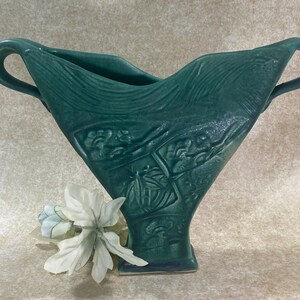 May include: A dark green ceramic vase with a wide, flared top and two side handles. The body features a textured, relief-molded pattern of rectangular panels containing floral motifs. A small, white artificial flower is placed at the base.