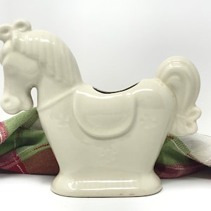 Könnte beinhalten: Weißer Keramik-Pferde-Pflanztopf mit Sattel und Schleife an der Mähne. Der Topf steht auf einem karierten Stoff.