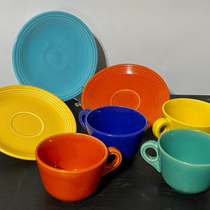 Könnte beinhalten: Ein Set aus Keramikgeschirr mit drei runden Untertassen in Türkis, Gelb und Orange sowie drei passenden Tassen in Orange, Blau und Gelb. Jedes Teil weist eine glatte, einfarbige Glasur und Details mit konzentrischen Ringen auf.