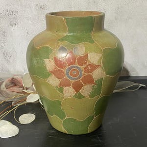 May include: A ceramic vase with a wide mouth and rounded body, featuring a matte finish in shades of olive green and tan. The surface is decorated with a repeating scalloped pattern and a central stylized flower motif in red, tan, and blue.