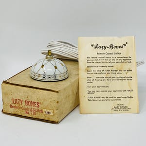 Puede incluir: Un interruptor de control remoto "Lazy Bones" vintage. El interruptor tiene una parte superior con estampado blanco y dorado y un cable blanco. Se encuentra en una caja de cartón con el logotipo "Lazy Bones" y una descripción del producto.