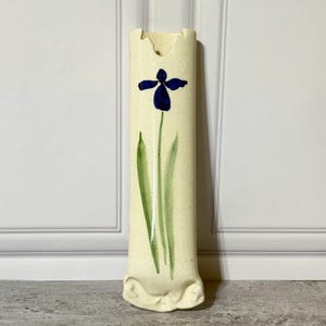 May include: A tall, cylindrical, off-white ceramic wall vase with a speckled finish. The front features a hand-painted design of a single dark blue flower with long, light green leaves extending downward. The top has a scalloped opening.