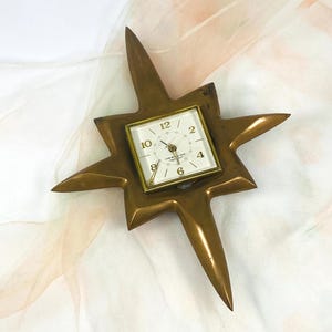 May include: A vintage starburst clock with a brass finish. The clock face is square with a white background and black numerals. The hands are gold-toned. The starburst design has pointed rays extending outwards.