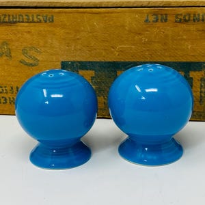 Puede incluir: Dos saleros y pimenteros esféricos de color azul brillante. Cada uno tiene una base redonda y pequeños agujeros en la parte superior. Los saleros y pimenteros son de cerámica con un acabado brillante. El fondo es una caja de madera.
