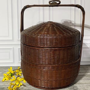 May include: A dark brown, round, three-tiered woven basket with a matching lid and a tall, arched handle. The structure is crafted from thin, tightly woven strips, featuring a smooth, polished finish and a secure, integrated carrying mechanism.