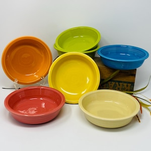 May include: A collection of six colorful ceramic bowls in shades of orange, red, yellow, green, blue, and cream. The bowls are round with a slightly flared rim, and are arranged on a white surface.