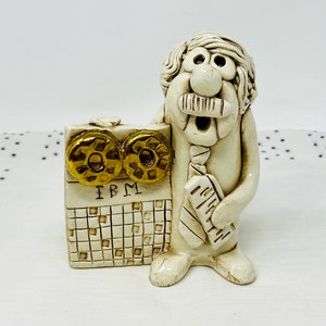 Puede incluir: Una figurita de cerámica blanca de un hombre con bigote y corbata, sosteniendo un objeto de cerámica blanca con las letras "IBM" y un patrón de cuadrícula. La figurita está de pie sobre una superficie blanca con un patrón de lunares azules.