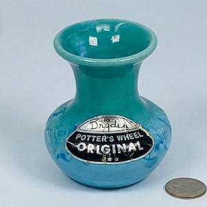 Könnte beinhalten: Kleine, türkisfarbene Vase mit rundem Boden und ausgestelltem Rand. Die Vase trägt ein Etikett mit der Aufschrift "Dryden Potter's Wheel Original". Eine Münze liegt daneben zum Größenvergleich.