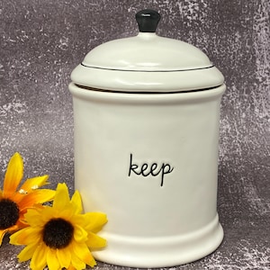 Puede incluir: Un bote de cerámica blanco con tapa y pomo negro. La palabra "keep" está escrita en escritura negra en la parte delantera. Dos girasoles amarillos con centros marrón oscuro están en primer plano.