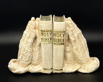 May include: Cream-coloured bookends featuring a pair of books with "HOLY BIBLE" text on the spines. The bookends are sculpted in the shape of praying hands, with a textured, aged appearance. The background is black.