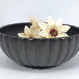 Könnte beinhalten: Eine dunkelgraue, gerippte Keramikschale gefüllt mit cremefarbenen Kunstblumen. Die Schale hat einen gewellten Rand und einen abgerundeten Boden. Die Blumen haben eine dunkelrote Mitte. Die Schale wird wahrscheinlich zu Dekorationszwecken verwendet.