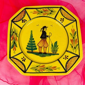 Op de afbeelding: Gele achthoekige decoratieve plaat met een handgeschilderd ontwerp. Het midden toont een figuur met een wandelstok, een boom en gebladerte. De rand heeft een zwart-groen bloemmotief. De plaat staat op een roze achtergrond.
