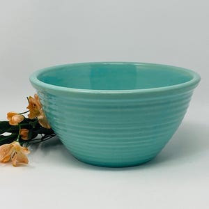 Peut inclure: Un saladier en céramique turquoise avec des détails côtelés horizontaux. Le bol a une forme arrondie et un intérieur lisse. Un petit bouquet de fleurs orange et jaunes repose à côté du bol, ajoutant une touche de couleur.