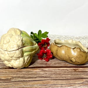 May include: A ceramic cauliflower-shaped lidded container with a cream and pale green finish, positioned next to a brown potato-shaped ceramic dish with a scalloped rim, both accented by two small red artificial flowers with green leaves.