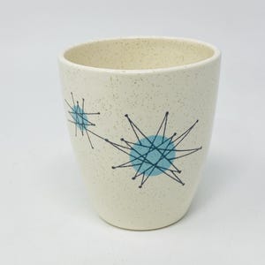 Puede incluir: Vaso de cerámica color crema con acabado moteado. Presenta dos diseños de estrellas turquesas con líneas negras. El diseño es de estilo moderno de mediados de siglo. Aproximadamente 10 cm de alto.