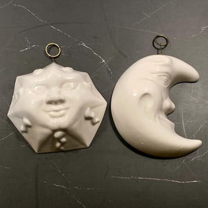 以下が含まれることがあります： 彫刻された顔が特徴の白い陶器製の壁掛け2点。1点は中央に顔がある幾何学的な太陽、もう1点は横顔が描かれた三日月です。それぞれのオーナメントの上部には、吊り下げるための小さな金属製のリングが付いています。