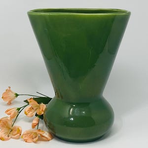 Puede incluir: Un jarrón de cerámica verde con una forma inusual de reloj de arena. El jarrón tiene una abertura ancha y cuadrada que se estrecha hasta una sección media bulbosa, luego se ensancha nuevamente en la base. Flores de color melocotón están dispuestas cerca del jarrón.