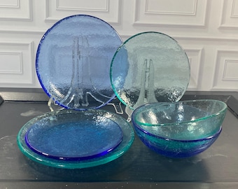 May include: A collection of glassware featuring two circular plates and two bowls in shades of blue and teal. The items have a textured, crackled appearance with translucent surfaces. One large blue plate and one smaller teal plate stand upright.
