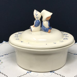 May include: White ceramic casserole dish with a lid. The lid features two small blue and white figurines of children sitting back to back. The dish is decorated with blue dots.