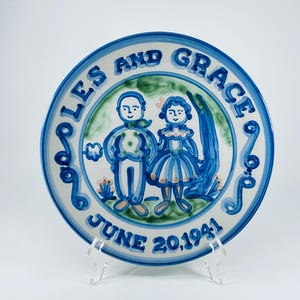 Puede incluir: Plato de cerámica pintado a mano con borde azul y blanco. El plato presenta una pareja con atuendo azul, con las palabras "JOLES AND GRACE" y "JUNE 20,1941" en letras azules. El plato se exhibe sobre un soporte transparente.