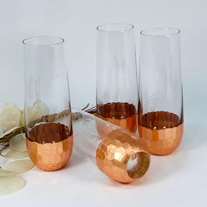 Può includere: Quattro flûte da champagne trasparenti con basi strutturate color rame. Una flûte è sdraiata su un lato. Le basi in rame hanno un aspetto di metallo martellato. Le flûte sono eleganti e adatte alle occasioni speciali.