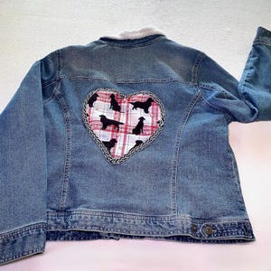 May include: A blue denim jacket featuring a white faux fur collar and a large heart-shaped patch on the back. The patch displays a red and white plaid pattern with black silhouettes of dogs and is bordered by a braided black and white trim.
