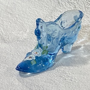 May include: A translucent blue glass shoe figurine featuring a molded texture and a hand-painted floral design on the toe. The decorative object has a pointed toe, a raised heel, and a scalloped top edge, showcasing a vibrant cobalt blue hue.