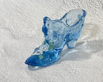 May include: A translucent blue glass shoe figurine featuring a molded texture and a hand-painted floral design on the toe. The decorative object has a pointed toe, a raised heel, and a scalloped top edge, showcasing a vibrant cobalt blue hue.