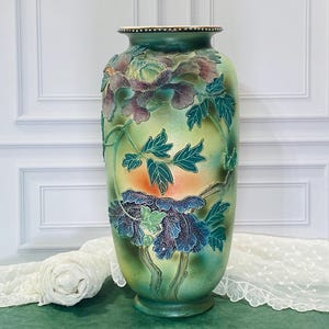May include: A tall ceramic vase featuring a gradient green and yellow background with raised, three-dimensional floral relief patterns. The flowers are colored in shades of purple and blue, accompanied by green leaves and stems on the exterior surface.
