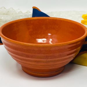 Puede incluir: Un cuenco de cerámica naranja vidriada con un borde ancho y un exterior acanalado. El cuenco se asienta sobre una base pequeña y tiene un interior brillante. Es un artículo de cocina.