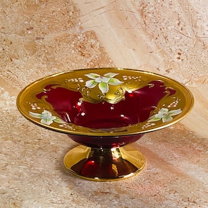 May include: A deep red glass pedestal bowl featuring a wide gold rim decorated with hand-painted white and green floral motifs. The bowl sits on a circular gold base, and the interior of the glass is a translucent ruby red color.