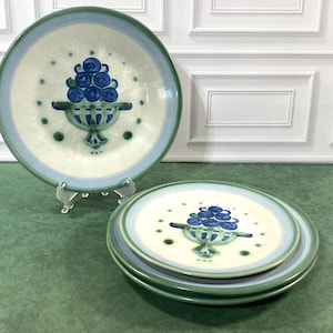 May include: A set of four round ceramic plates featuring a hand-painted design of a blue fruit-filled urn centered on a cream-colored background. Each plate has a wide light blue and sage green border with small green dots surrounding the central motif.