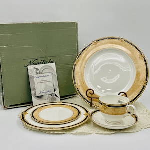 Könnte beinhalten: Ein Noritake-Geschirr-Set mit goldenen und blauen Verzierungen. Das Set enthält einen Speiseteller, einen Salatteller, eine Teetasse und eine Untertasse. Das Set ist in einer grünen Box mit dem Noritake-Logo verpackt.