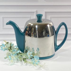 Puede incluir: Una tetera con cuerpo de metal plateado pulido y asa, pico, pomo de tapa y base de color verde azulado. La tetera está decorada con un grupo de flores artificiales de color azul claro situadas en la base frontal del recipiente.