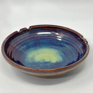 May include: A handmade ceramic ashtray with a glossy finish. The bowl features a blend of blue, purple, and yellow hues, with a brown rim and built-in rests for cigarettes. The interior has a swirling pattern, adding to its unique design.