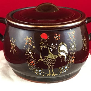 May include: A brown ceramic crock pot with a lid features a rooster design with gold and red accents. The rooster is surrounded by white and orange flowers.