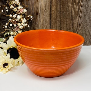 Könnte beinhalten: Eine orangefarbene Keramikschüssel mit gerippter Außenseite. Die Schüssel ist rund und hat eine weite Öffnung. Der Hintergrund umfasst eine Holzoberfläche und Blumendekorationen. Diese Schüssel eignet sich zum Servieren von Speisen.