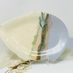 Può includere: Piatto da portata in ceramica a forma di pesce. Il piatto è diviso in due sezioni, con un lato color crema e un lato azzurro. Strisce verticali verdi, blu e pesca aggiungono colore. Il piatto è sostenuto da un supporto trasparente.