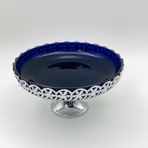 May include: A cobalt blue glass bowl with a smooth interior, set upon a silver-toned metal pedestal base. The rim of the bowl is encircled by a decorative silver-toned metal band featuring a repeating openwork floral or circular pattern.