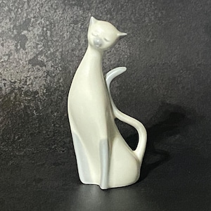 May include: A smooth, off-white ceramic cat figurine featuring an elongated neck, a pointed head with closed eyes, and a curved tail that arches upward. The surface has a glossy finish with subtle light gray shading on the lower legs and tail.