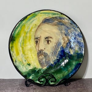 May include: A round ceramic plate featuring a hand-painted portrait of a bearded person with blue hair. The background consists of swirling yellow, green, and blue brushstrokes. The plate has a dark rim and rests on a black metal display stand.