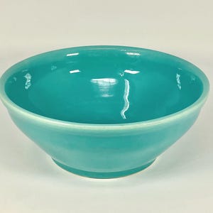 May include: A turquoise ceramic bowl with a glossy finish. The bowl has a rounded shape with a slightly flared rim. The interior and exterior are the same color, and the bowl sits on a white surface.