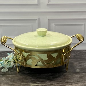 May include: A round, light green speckled ceramic casserole dish with a matching lid, set inside a decorative gold-toned metal stand. The stand features intricate cut-out patterns and two side handles with a coiled wire design.