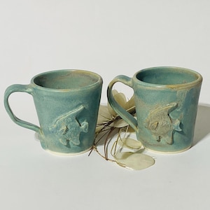 May include: Two handmade ceramic mugs in a muted teal glaze, each featuring a raised, embossed angelfish design on the front. The mugs have a slightly tapered shape, a sturdy handle on the side, and a smooth, glossy finish with subtle color variations.