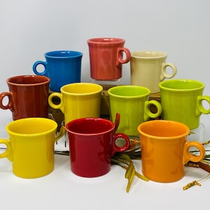 Puede incluir: Una colección de diez tazas de cerámica en varios colores, incluyendo rojo, amarillo, azul y verde. Cada taza tiene un asa redondeada y una forma cilíndrica. Las tazas están dispuestas sobre una superficie blanca.