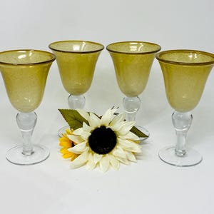 May include: Four amber-colored wine glasses with clear stems and bases. The glasses have a speckled texture and a gold rim. A sunflower with white and yellow petals sits in front of the glasses.