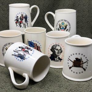 May include: Set of six white ceramic mugs with patriotic designs. Each mug features a different image related to American history, including the Great Seal of the United States, George Washington, and a drummer boy.