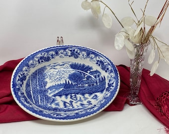 May include: A white oval serving bowl with a blue and white floral pattern. The bowl features a blue and white landscape scene with a house, trees, and mountains.